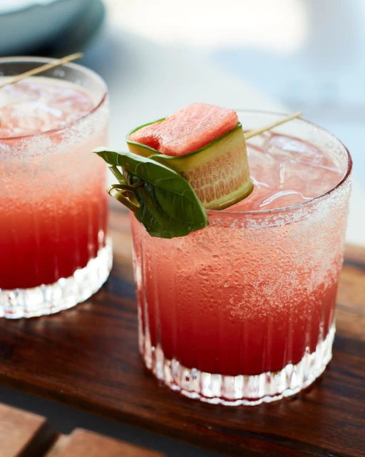 Two refreshing cocktails with ice, garnished with watermelon, cucumber, and a green leaf, served on a wooden tray.