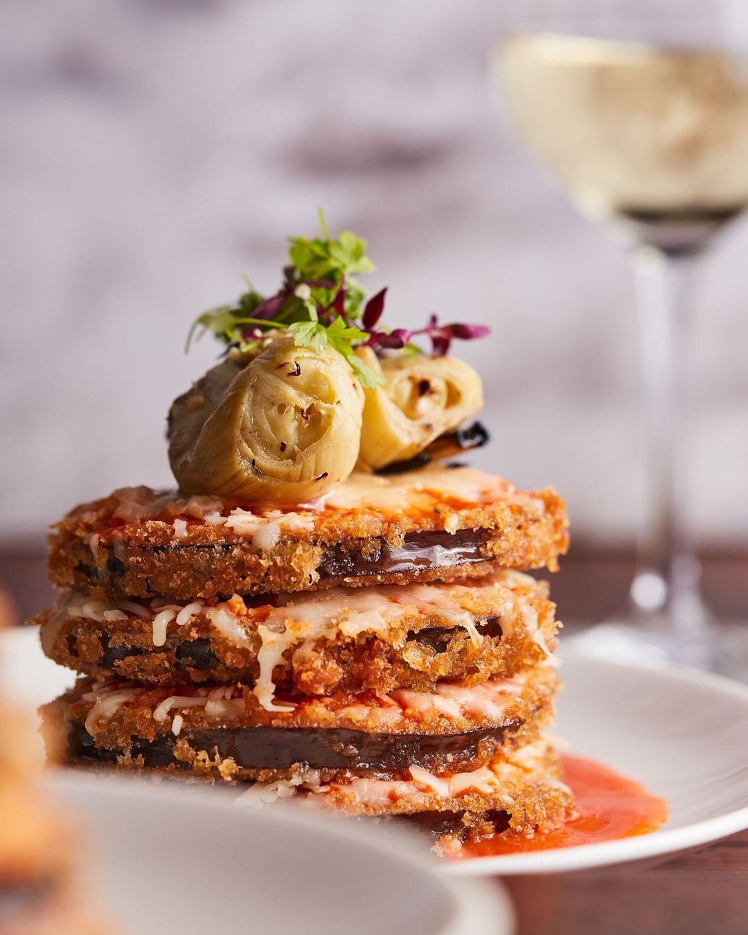 Stack of eggplant parmesan topped with artichoke hearts and herbs, served on a white plate with a blurry glass of white wine.