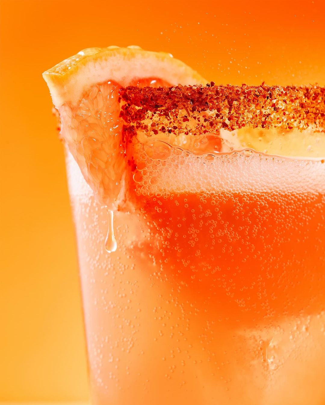 Close-up of a vibrant coral-colored cocktail in a glass, garnished with a grapefruit slice and a spiced rim against a warm orange backdrop.