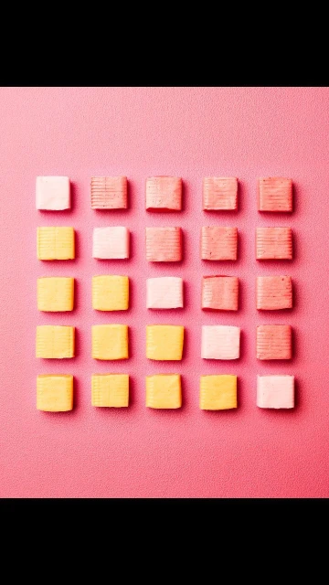 Overhead shot of pink, yellow, and white square confections in neat rows on a pink backdrop. Candies are arranged in a grid pattern.