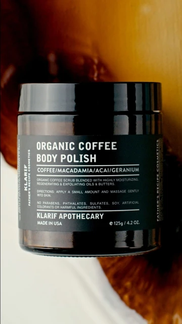 A jar of Klarif Organic Coffee Body Polish on a smooth surface with a blurred brown and tan background creates a natural beauty product image.