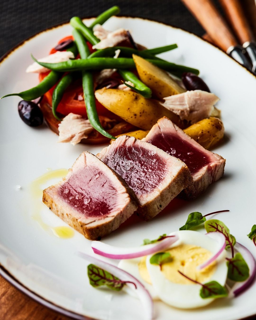 Plated seared tuna salad featuring rare tuna slices, green beans, tomatoes, potatoes, hard boiled eggs, and red onion.