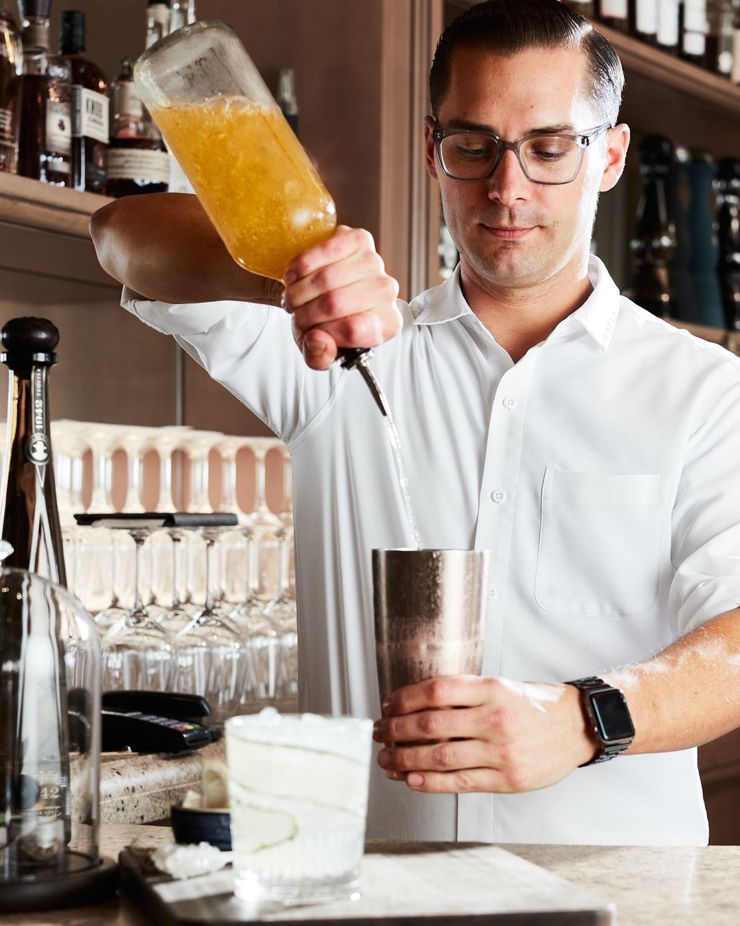 Adult man bartender preparing a cocktail. He is pouring liquid into a metal shaker, and a finished cocktail sits in the foreground.