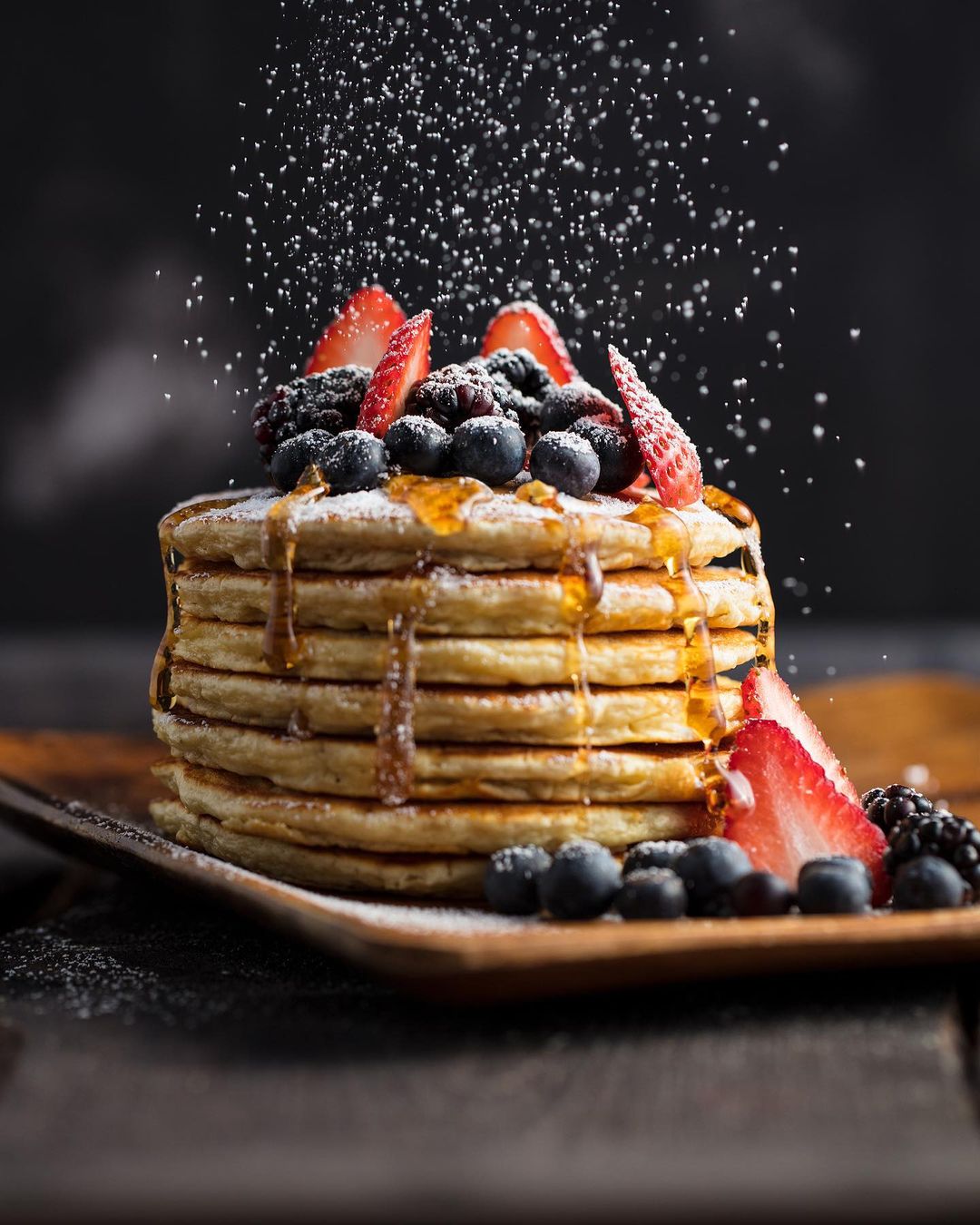 Elevated view of a stack of pancakes drizzled with syrup and topped with mixed berries, set against a dark backdrop with sugar sprinkling.