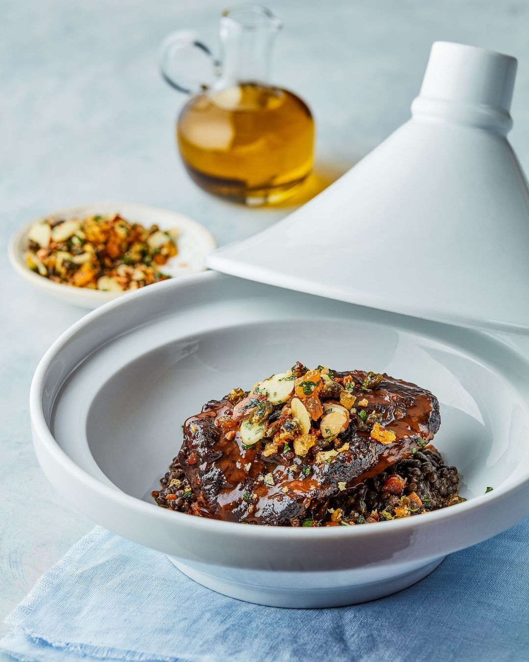 A close-up studio shot showcases a gourmet tagine dish with a white ceramic lid, served with lentils and garnished with nuts and herbs.