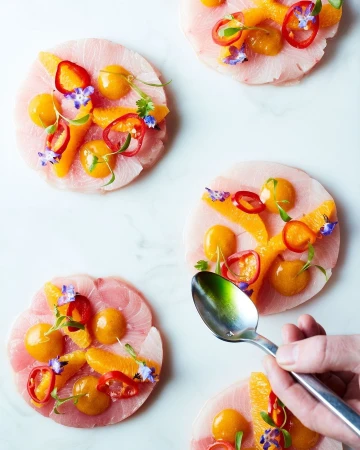 Overhead shot of raw fish slices arranged with orange segments, red pepper rings, edible flowers, and golden tomatoes, being garnished with a spoon.