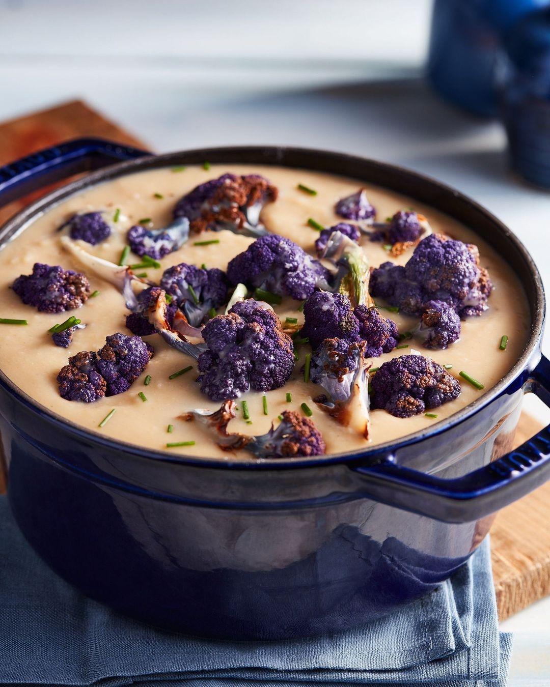 Overhead shot of vibrant purple cauliflower florets in creamy soup, presented in a dark blue pot on a rustic wooden board, offering a visually appealing food image.