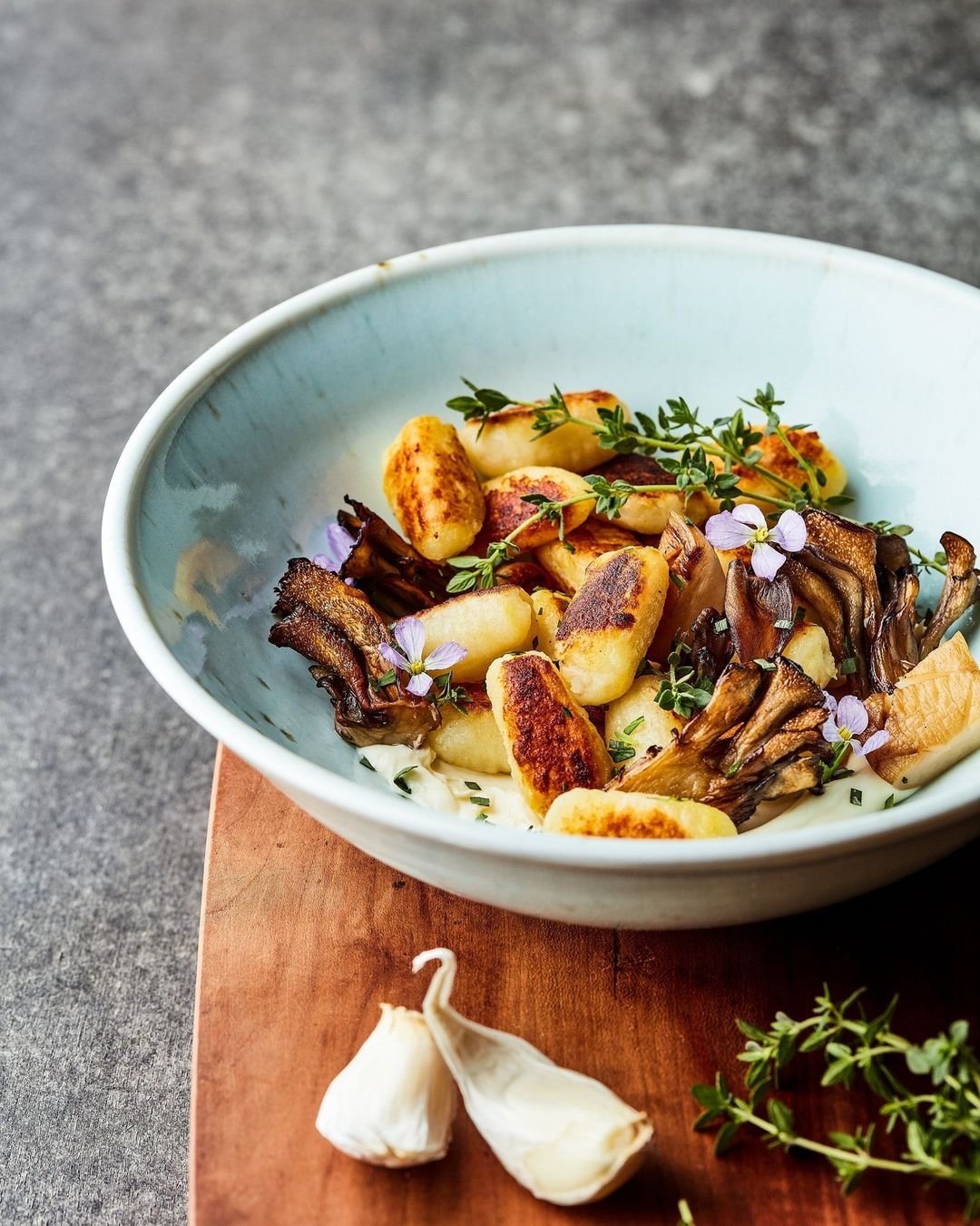 Close-up of gnocchi dish featuring pan-fried gnocchi with mushrooms, thyme sprigs, and garlic cloves on a rustic wooden board.