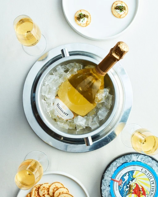 Overhead shot of chilled champagne in an ice bucket, surrounded by plates of appetizers and flutes of champagne on a white tablecloth.