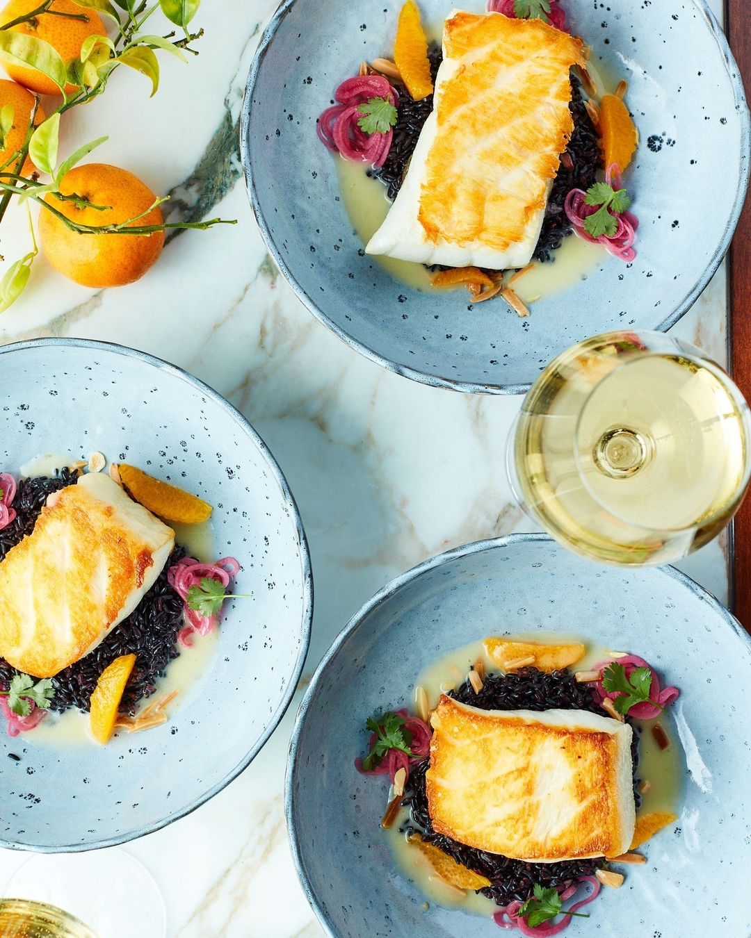 Overhead shot of three plates of grilled fish over black rice with sliced orange, red onion, and cilantro, served with white wine.
