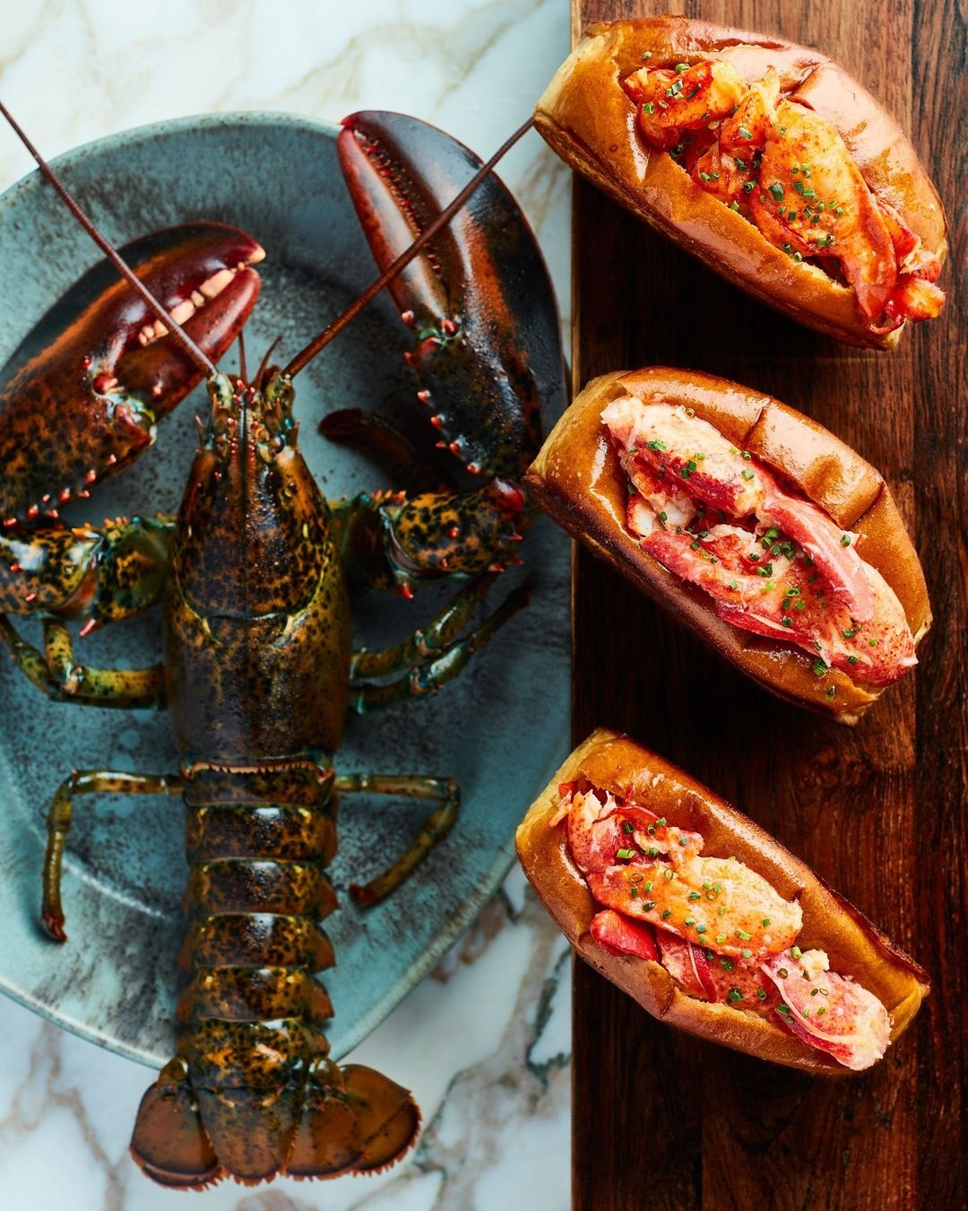 Overhead shot of a cooked lobster on a grey plate and three lobster rolls on a wood board, showing a culinary seafood dish.