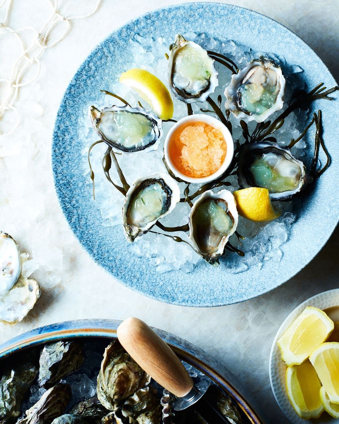 Overhead shot of fresh oysters served on ice with lemon wedges and orange dipping sauce, arranged on blue ceramic plates.