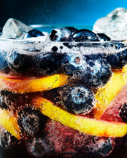 Close-up of a refreshing beverage with blueberries, sliced oranges, ice cubes, and bubbles against a dark blue background.