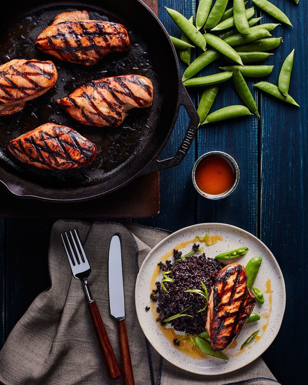 Overhead shot of cooked chicken breasts with grill marks on a cast iron skillet and plate, accompanied by black rice and sugar snap peas.