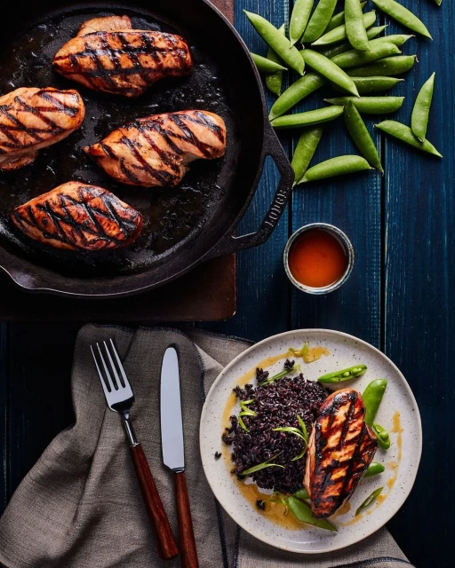 Overhead shot of cooked chicken breasts with grill marks on a cast iron skillet and plate, accompanied by black rice and sugar snap peas.