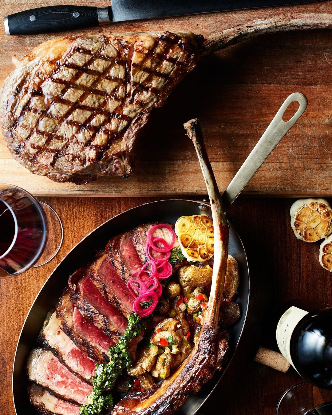 Overhead shot of grilled tomahawk steak sliced in a metal pan with potatoes and garlic, next to a bottle of red wine and a knife.