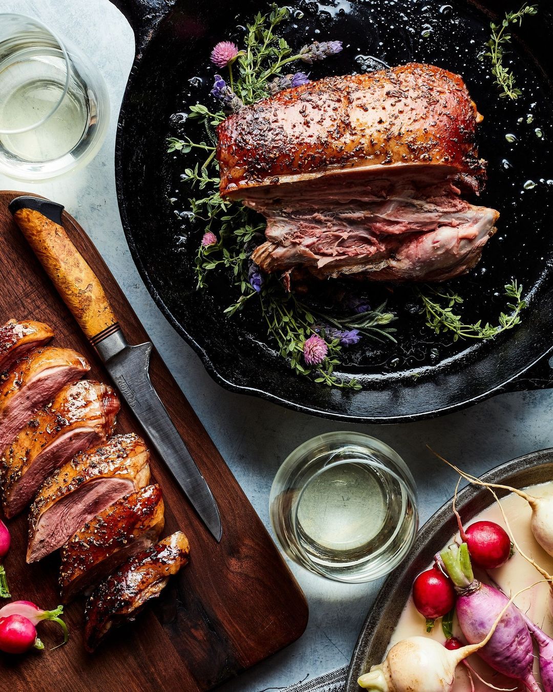 Overhead shot of sliced and whole duck meat on a cast iron pan and wooden board, accompanied by radishes and white wine.