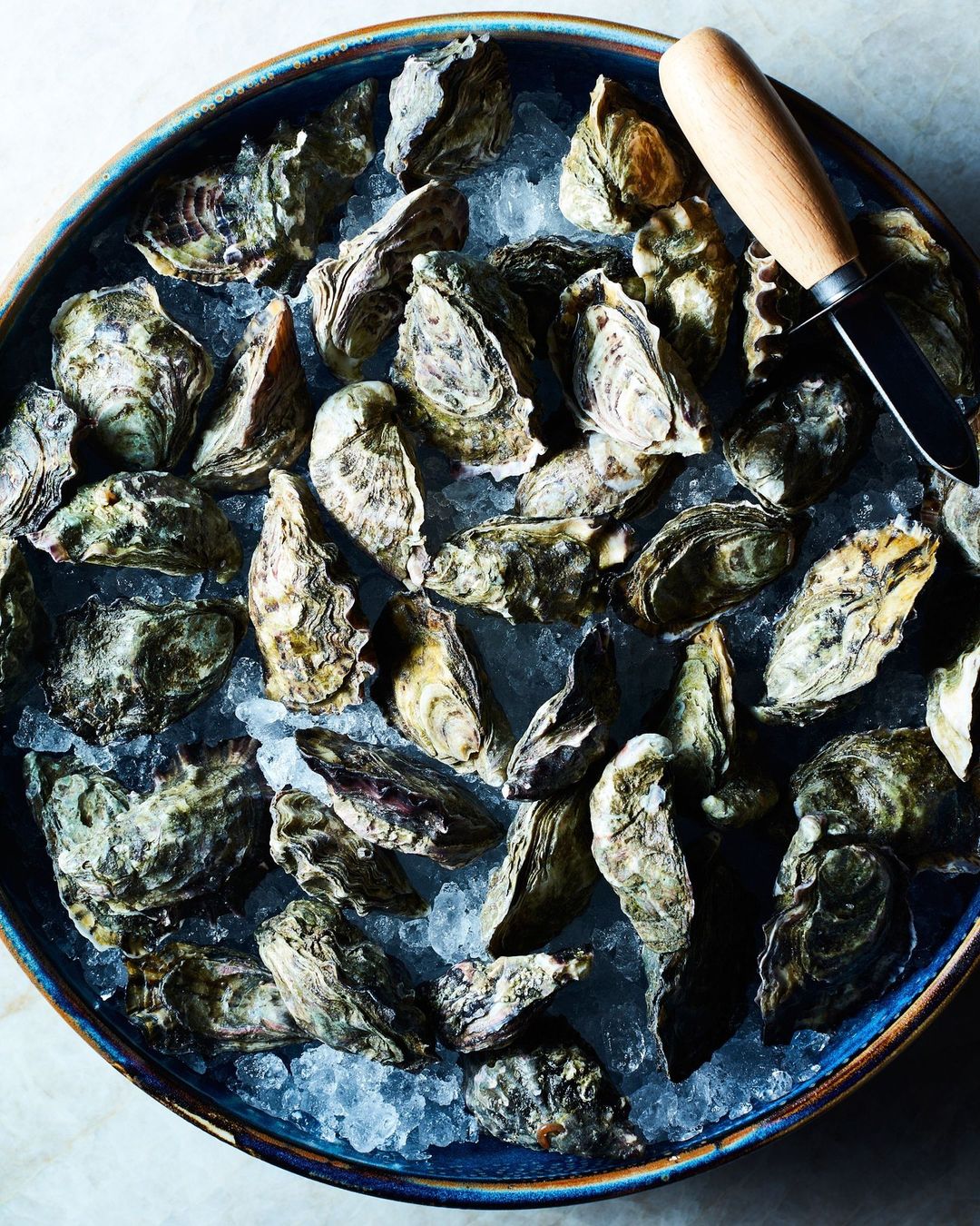Overhead shot of fresh oysters on a blue plate with crushed ice and an oyster knife, showcasing a seafood culinary experience.