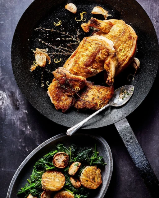 Overhead shot of roasted chicken in skillet with herbs and lemon zest, accompanied by sauteed spinach and roasted garlic on dark surface.