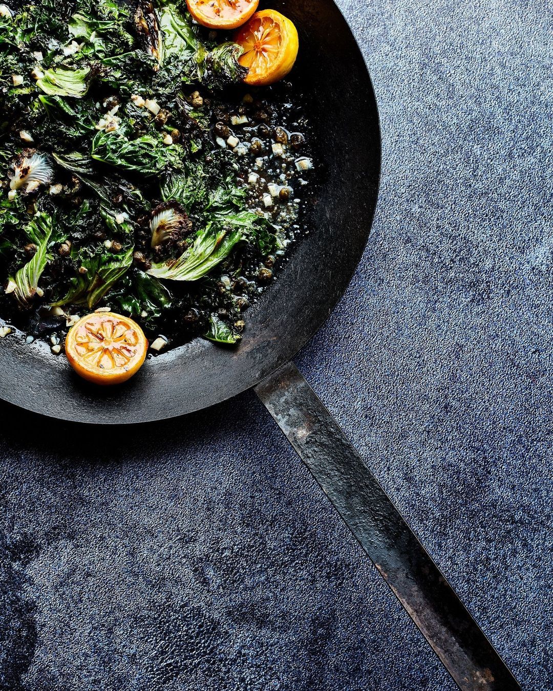 Overhead shot of sauteed greens with lemon wedges in a black pan, set against a textured dark gray background.