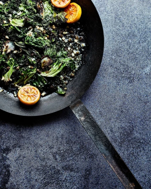 Overhead shot of sauteed greens with lemon wedges in a black pan, set against a textured dark gray background.
