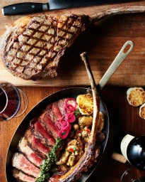 Overhead food photography by Skyler Burt showing sliced tomahawk steak with chimichurri and roasted potatoes in a skillet on a wooden table.