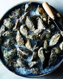 Top-down view of fresh raw oysters nestled on crushed ice inside a dark blue ceramic bowl with an oyster shucking knife resting on top.