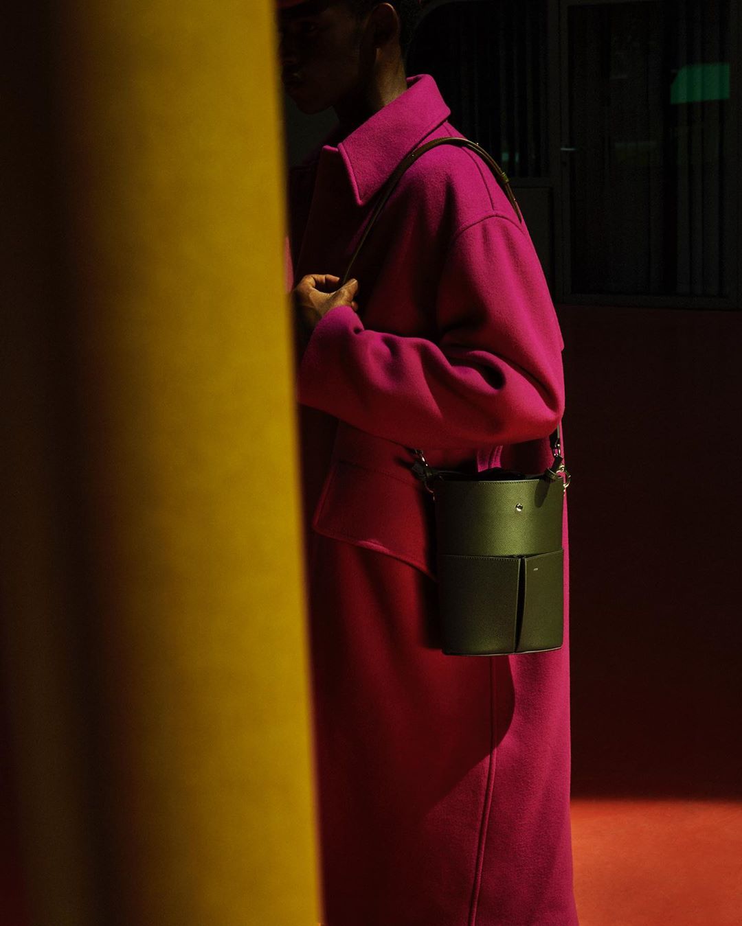 Woman wearing magenta coat with dark green cylindrical shoulder bag standing near a gold column in a dimly lit room.