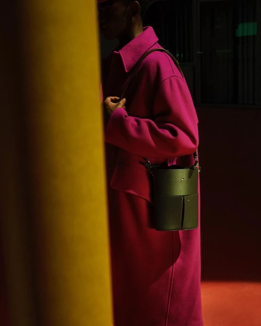 Woman wearing magenta coat with dark green cylindrical shoulder bag standing near a gold column in a dimly lit room.