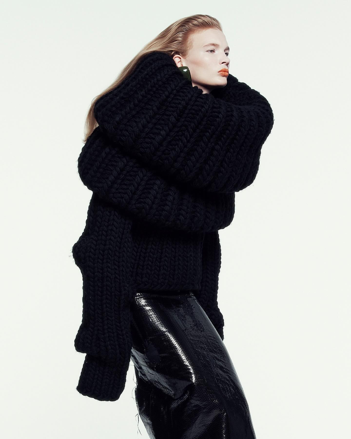 Fashion shot of a blonde adult female model wearing a chunky black knit sweater and shiny black pants against a white backdrop.