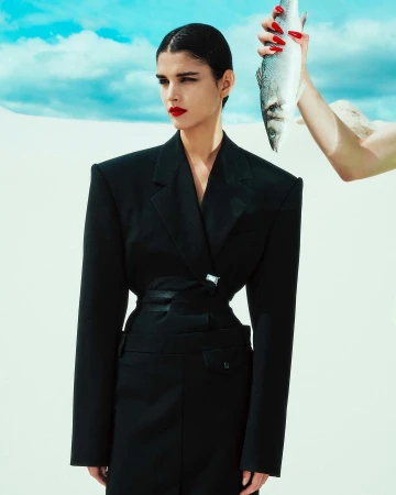 Woman in black suit poses in bright outdoor setting with fish held by another person, creating a surreal and fashion-forward image.
