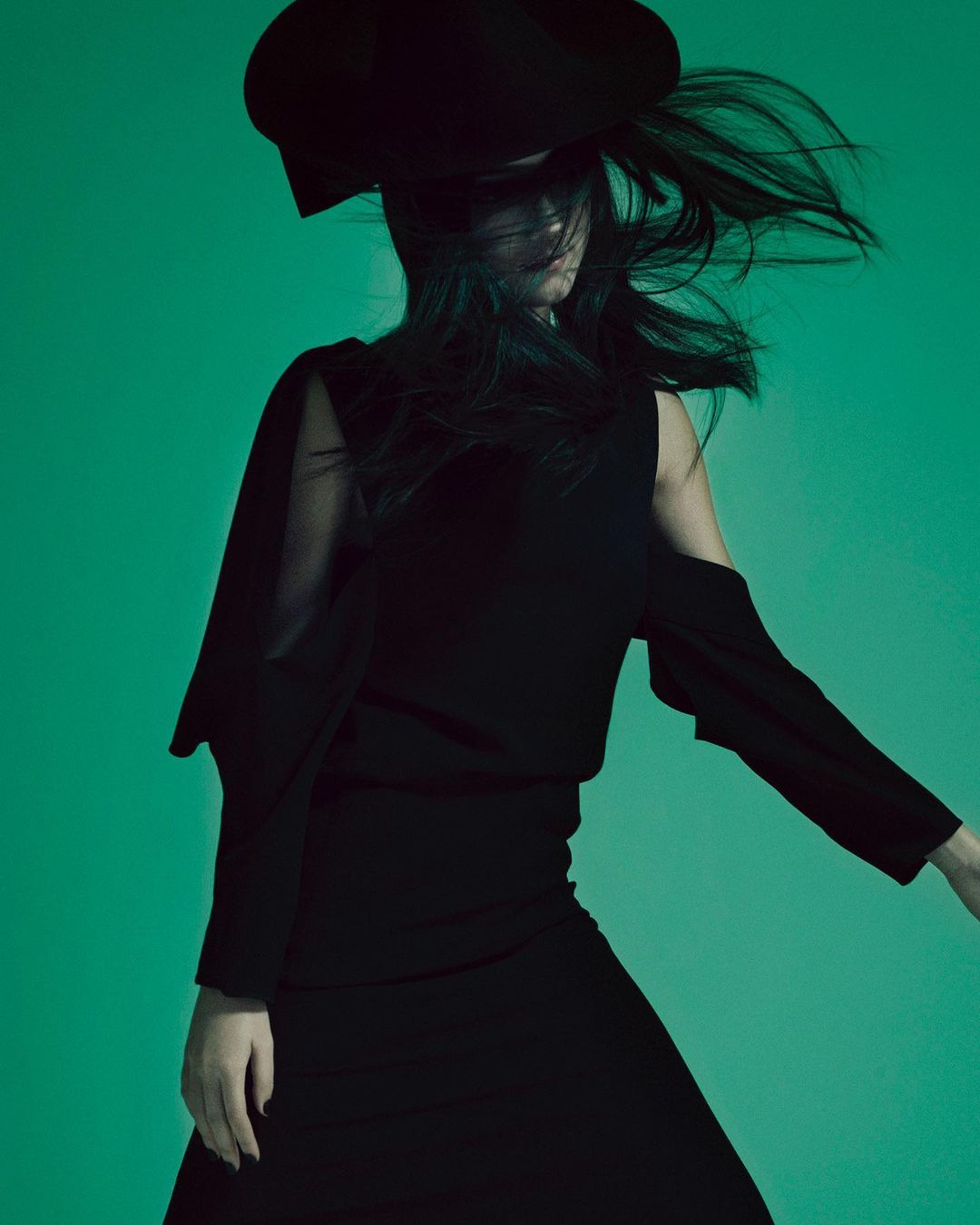 A woman in a black dress and a large hat poses against a green backdrop, capturing a fashion-forward and stylish portrait. 