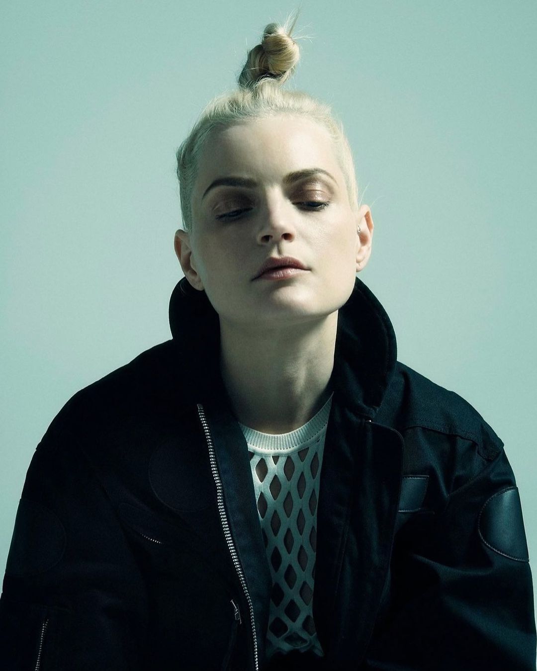 Close-up studio shot of a fair-skinned woman with a blonde topknot, wearing a black jacket and mesh top against a light blue background.