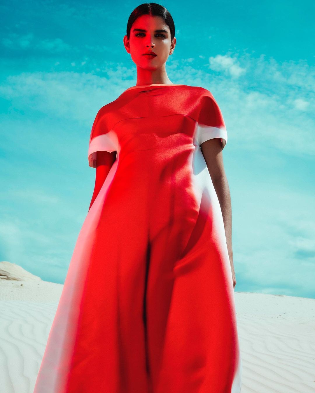 Woman in a red and white dress stands in a bright desert against a light blue sky with scattered clouds in a fashion editorial shot.