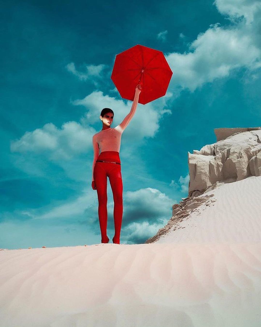 A female fashion model wearing red leggings stands in a desert landscape holding a red umbrella against a blue sky with white clouds.