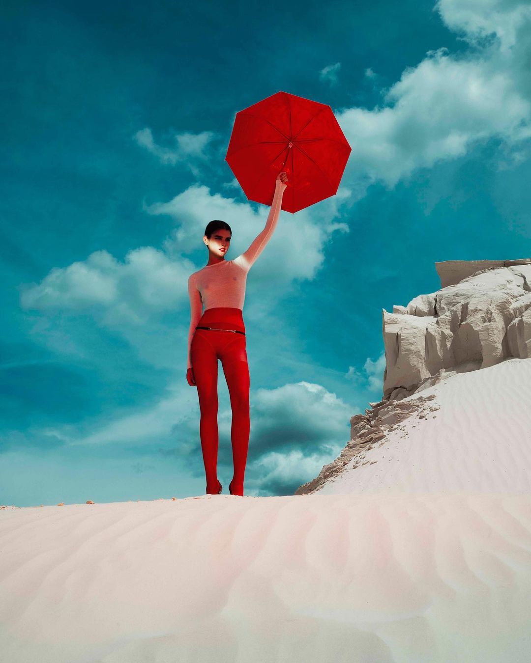 Fashion model in red pants stands on a sand dune, holding a red umbrella against a blue sky with light clouds. Outdoor scene.