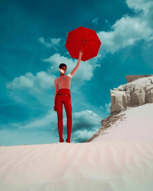 Fashion model in red pants stands on a sand dune, holding a red umbrella against a blue sky with light clouds. Outdoor scene.