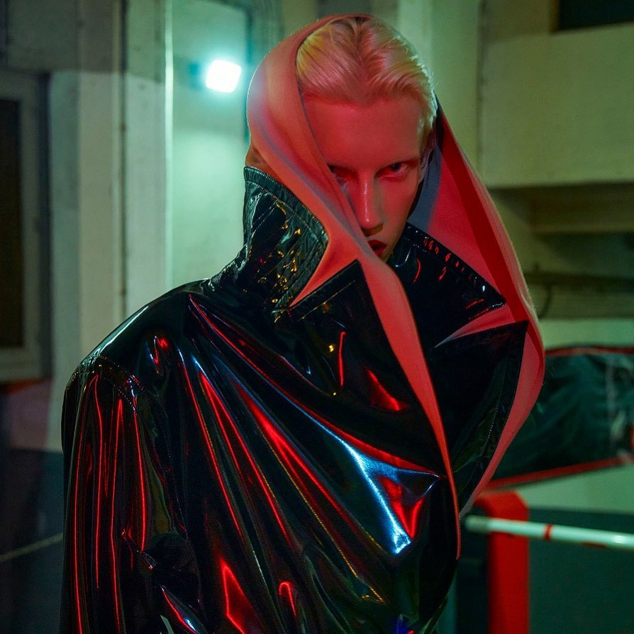 Androgynous individual in a glossy black jacket with a pink hood, striking a pose with dramatic lighting in an indoor environment.