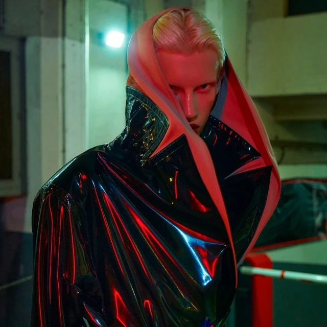 Androgynous individual in a glossy black jacket with a pink hood, striking a pose with dramatic lighting in an indoor environment.