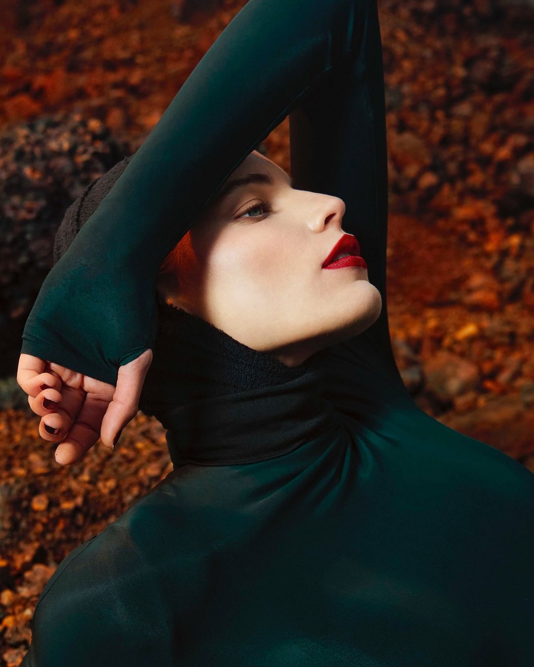 Woman in dark green turtleneck poses against a rocky, reddish-brown backdrop, face tilted upwards, arms above head, dark nail polish.
