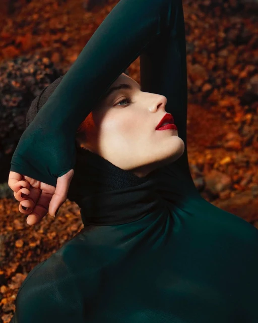 Woman in dark green turtleneck poses against a rocky, reddish-brown backdrop, face tilted upwards, arms above head, dark nail polish.
