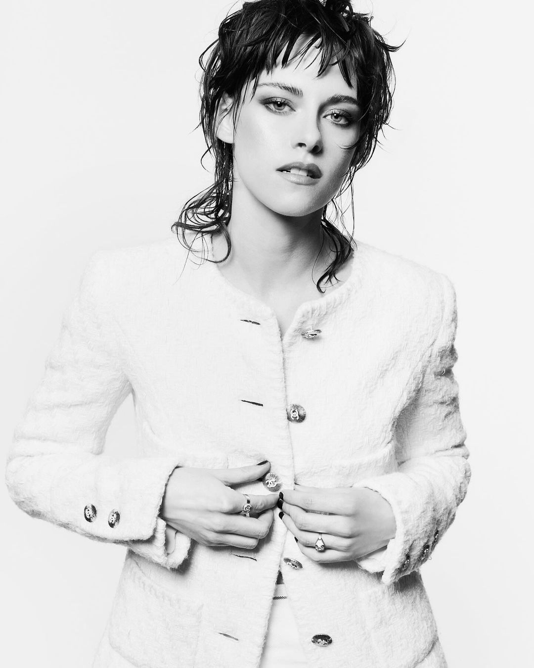 Black and white portrait of a woman with wet hair wearing a textured tweed jacket buttoning up, against a plain white background.