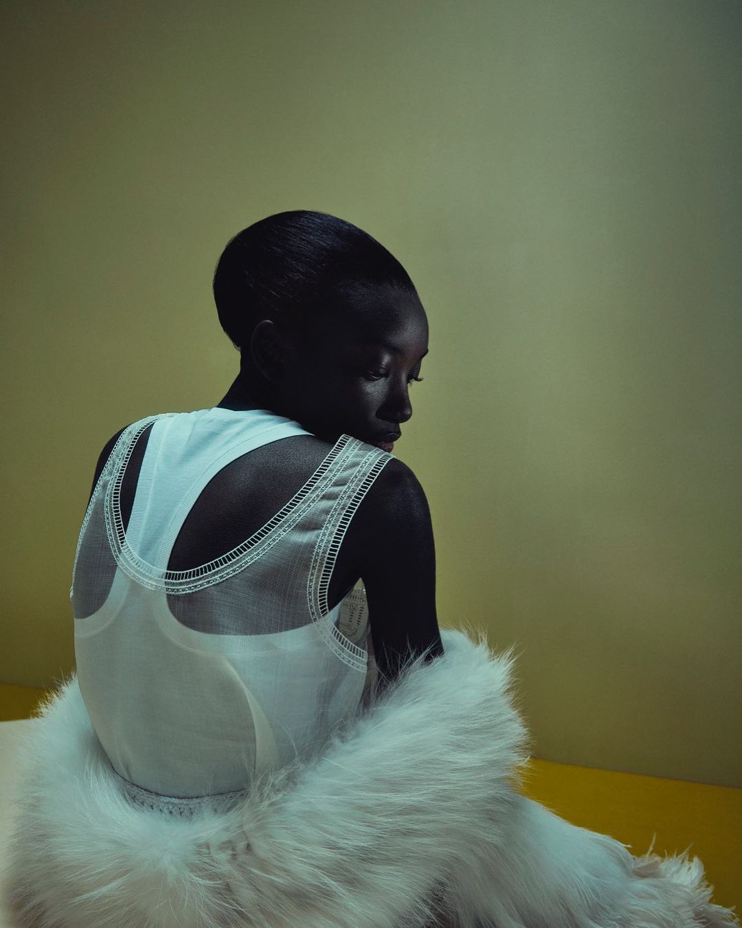 An African-American woman is seen from the rear wearing a white top and a fluffy white fur shawl in a studio setting with yellow background.