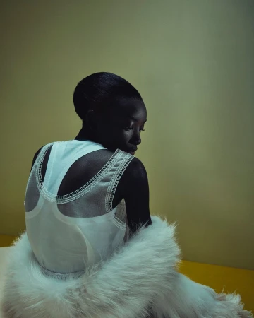 An African-American woman is seen from the rear wearing a white top and a fluffy white fur shawl in a studio setting with yellow background.