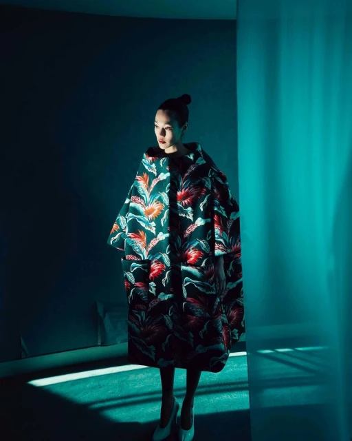 A stylish woman in a floral coat poses in a dimly lit blue room, highlighted by beams of light, showcasing a blend of fashion and modern photography.