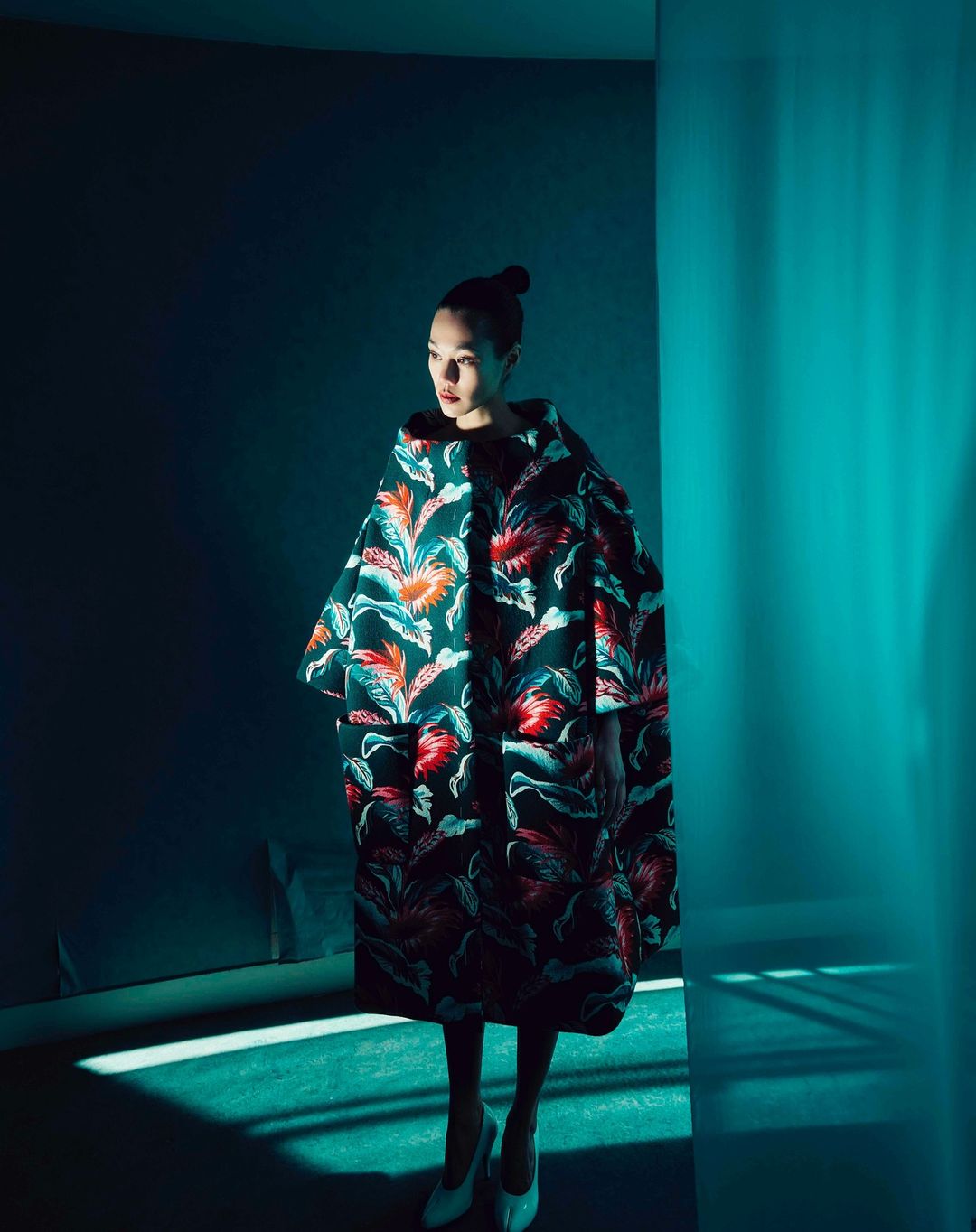Elegant woman in a floral coat poses in a minimalist room with teal lighting, showcasing a unique fashion statement.
