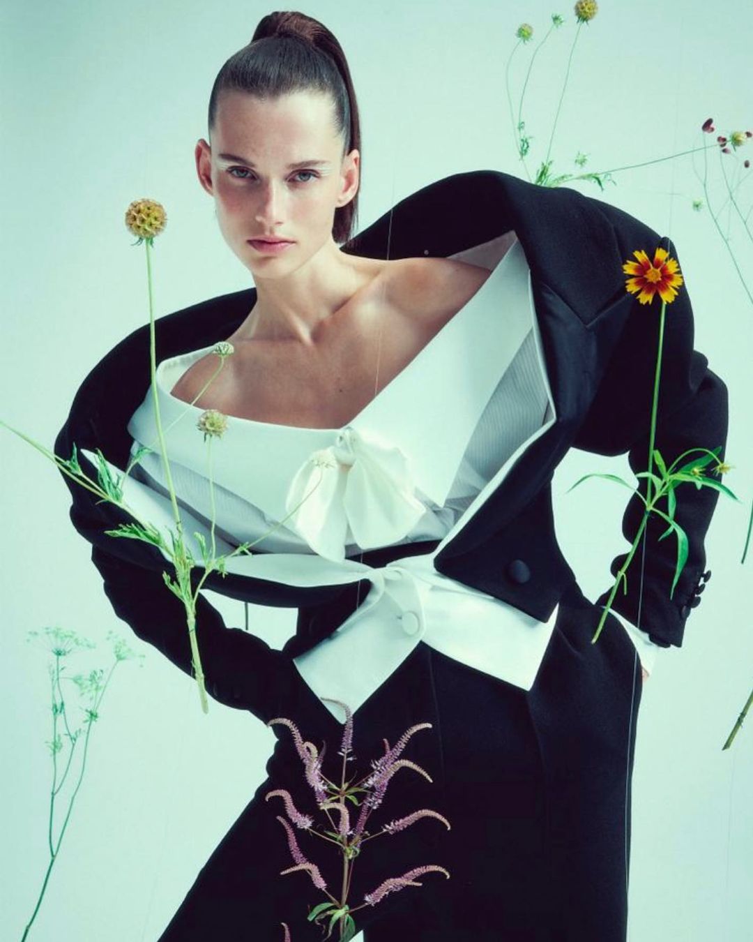 Fashion shot of a woman wearing a black suit jacket and white top, surrounded by flowers, against a light blue background in studio lighting.