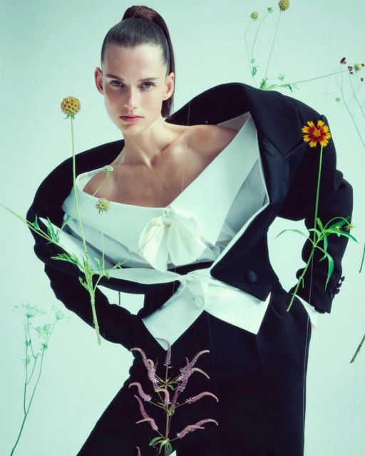 Fashion shot of a woman wearing a black suit jacket and white top, surrounded by flowers, against a light blue background in studio lighting.