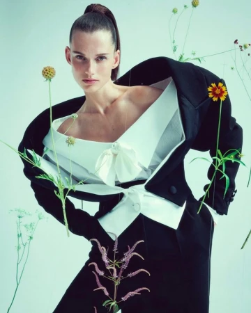 Fashion shot of a woman wearing a black suit jacket and white top, surrounded by flowers, against a light blue background in studio lighting.