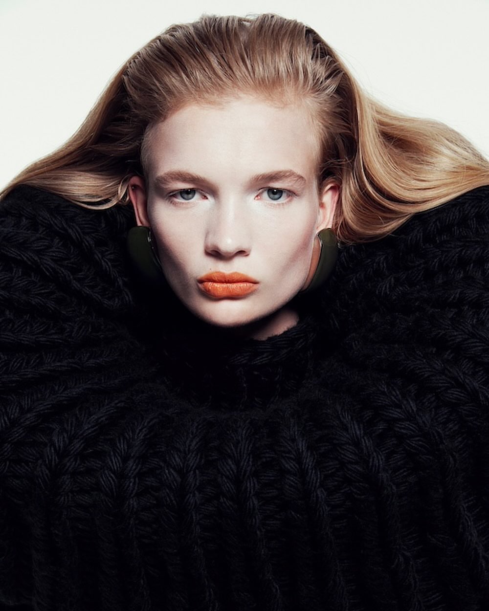 Portrait of a blonde woman wearing a chunky black knit sweater and bright orange lipstick against a white background.